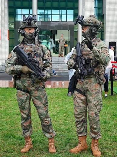 Two special operations soldiers standing on grass in full kit with helmet-mounted night-vision devices, rifles held at the ready outside a modern building