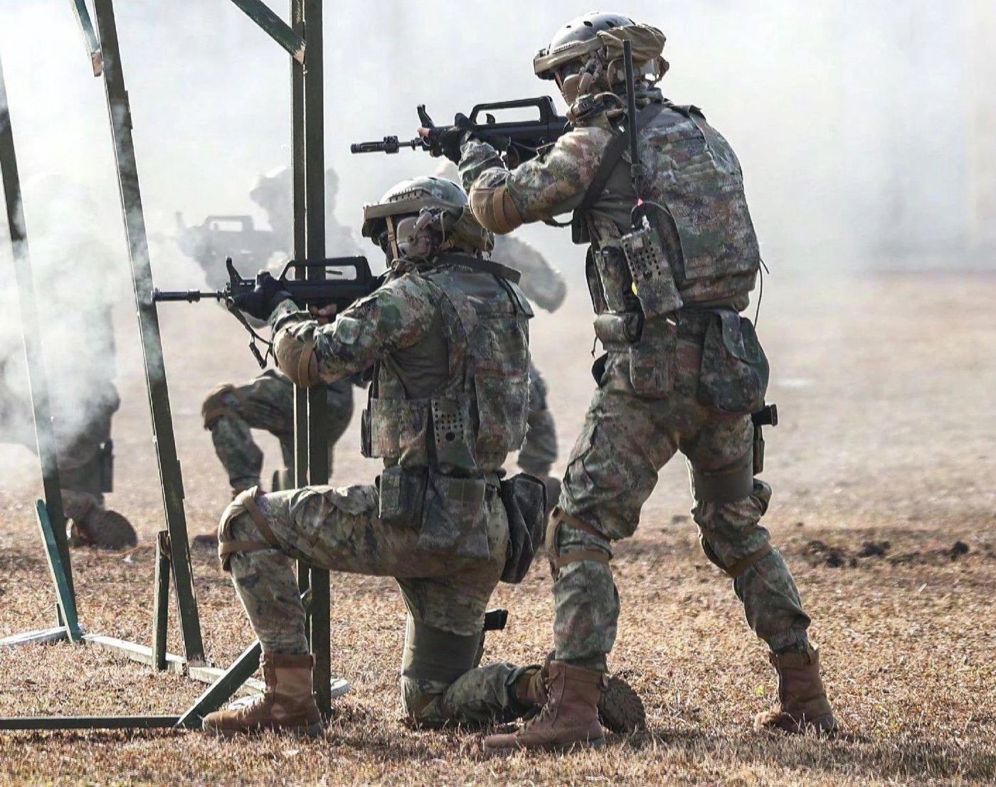 Two PLA special operations soldiers in digital camouflage firing rifles from behind a training barricade during a live-fire movement drill