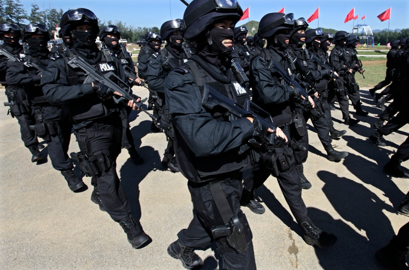 A column of tactical response operators in black uniforms, helmets and goggles marching in formation on a sunny training ground